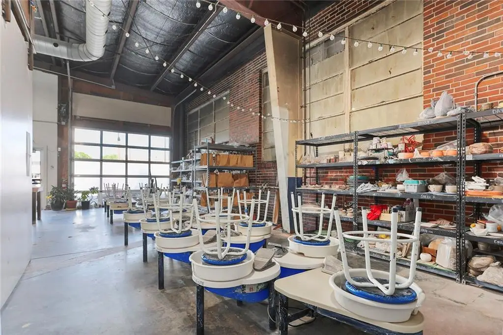 Interior of a warehouse with chairs stacked upside down on tables, shelves with various items, and large windows at the back.