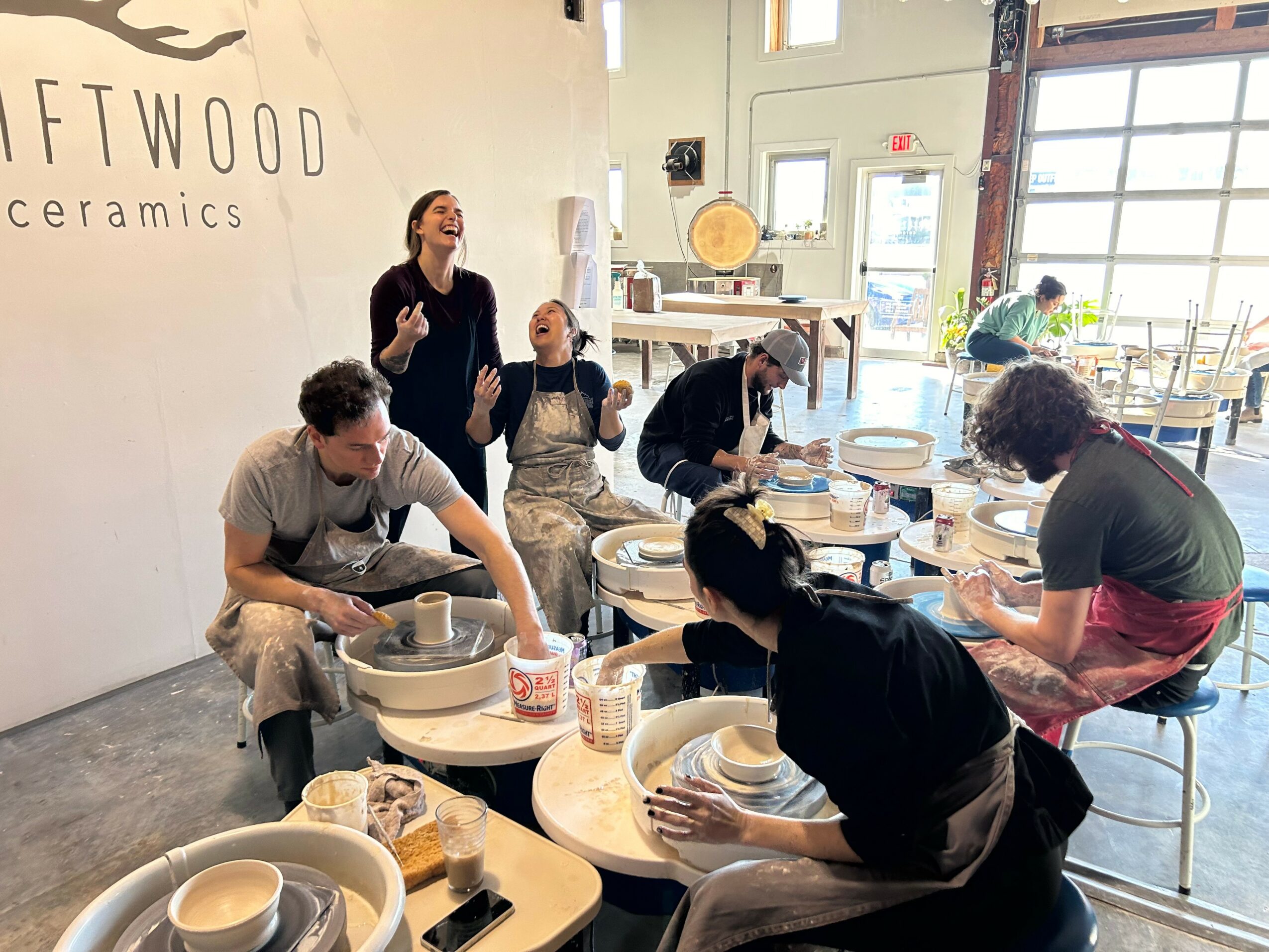 People working on pottery wheels in a ceramics studio with large windows and a sign reading 'FTWOOD ceramics'.
