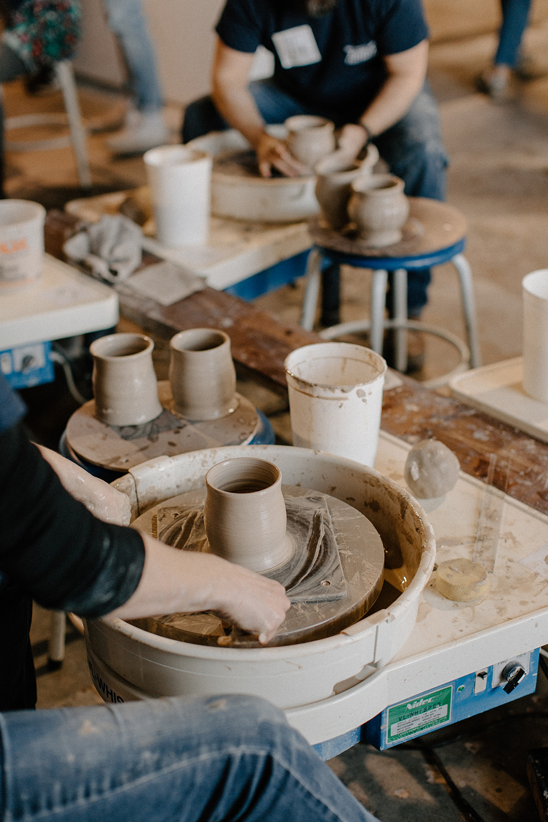 People working on pottery wheel creating clay cups in a workshop, with pottery tools and supplies on tables.