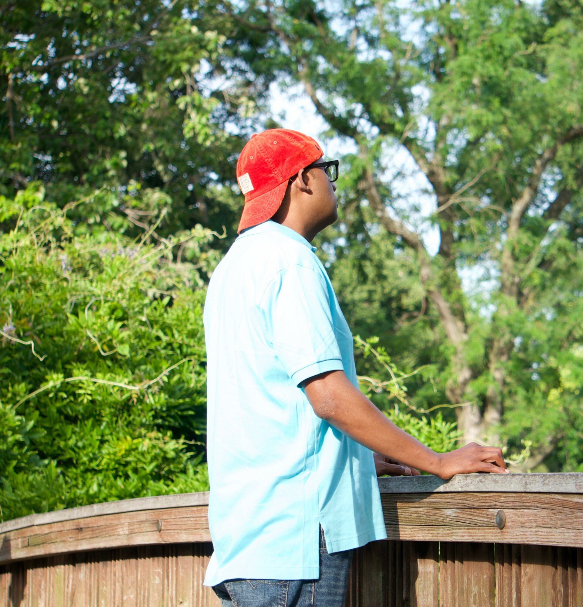 Person wearing a red cap, glasses, and a light blue shirt, standing outdoors near a wooden railing with green trees in background.