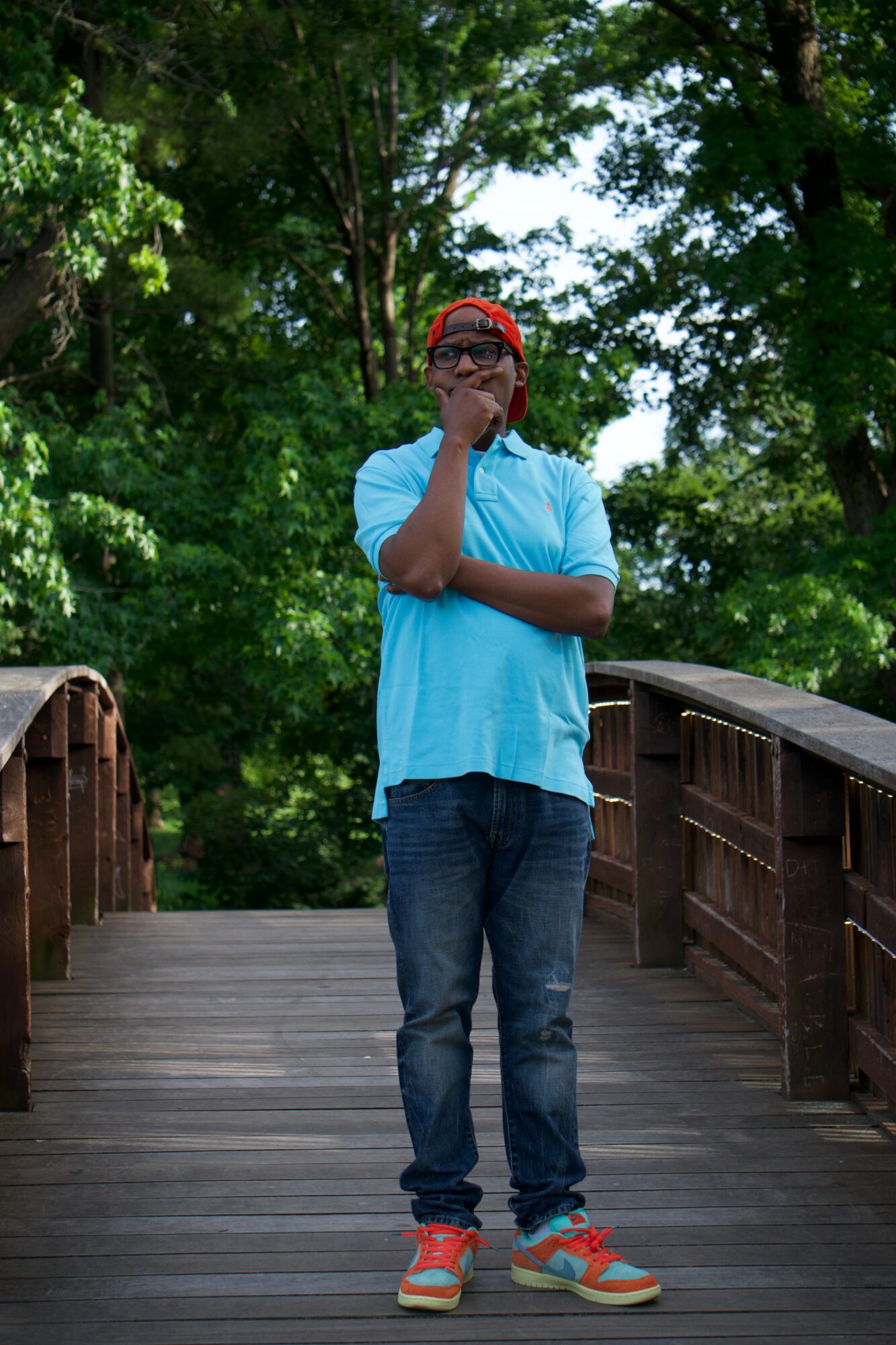 Person standing on a wooden bridge with trees in the background, wearing a light blue shirt, jeans, and colorful sneakers, holding a hand to face.