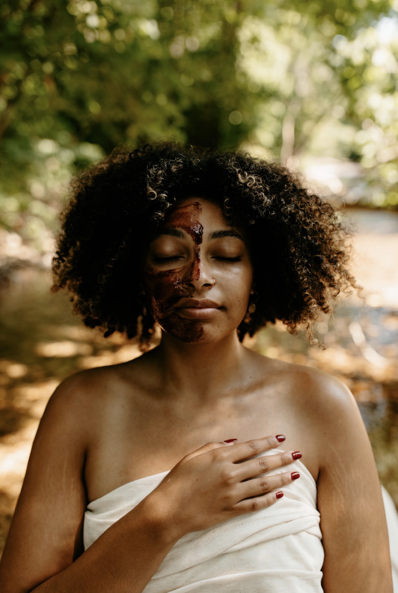 Young woman with curly hair, eyes closed, holding her chest with one hand outdoors, sunlight filtering through trees.