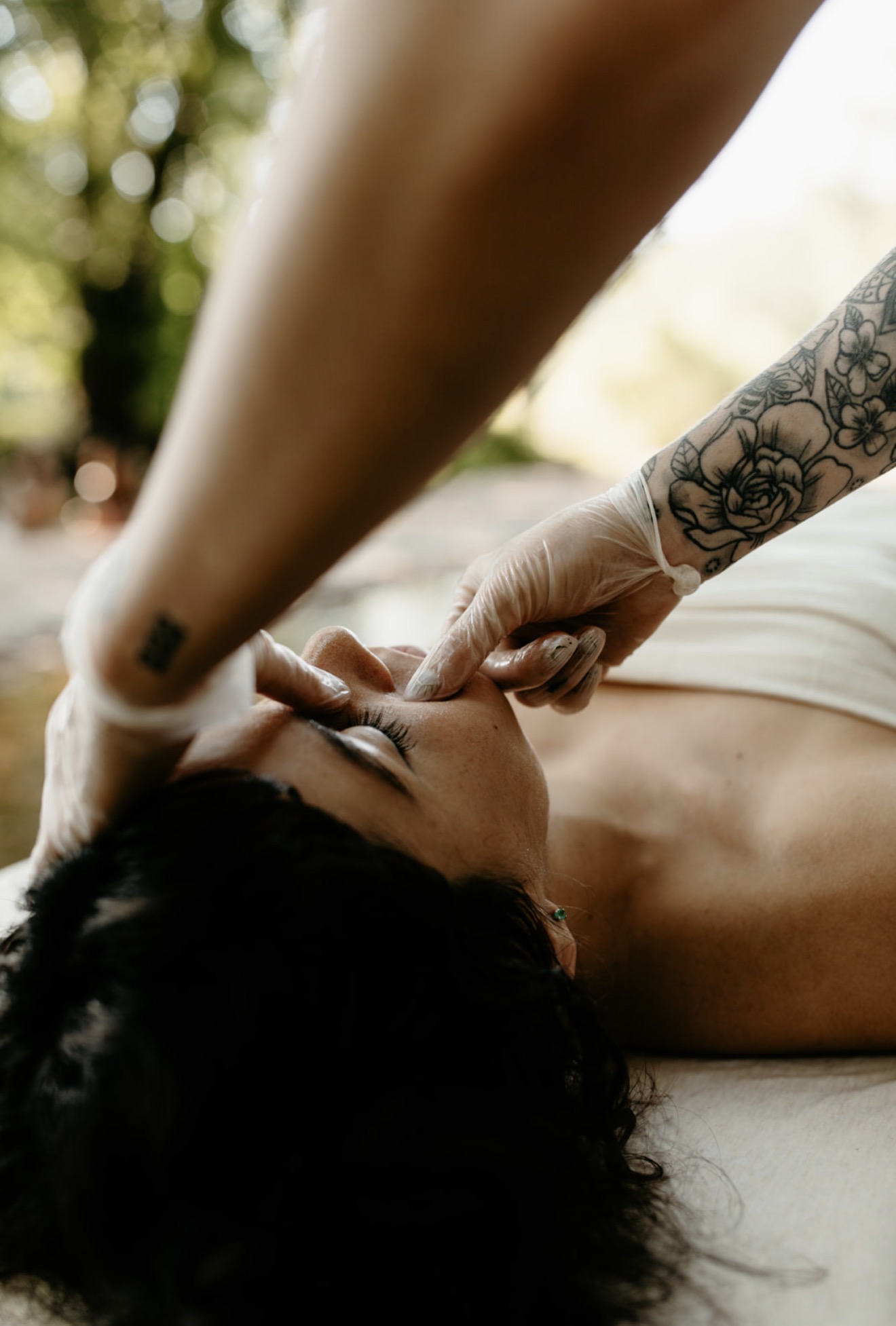 Person lying down receiving a massage outdoors, with a tattooed arm visible, and a blurred natural background.