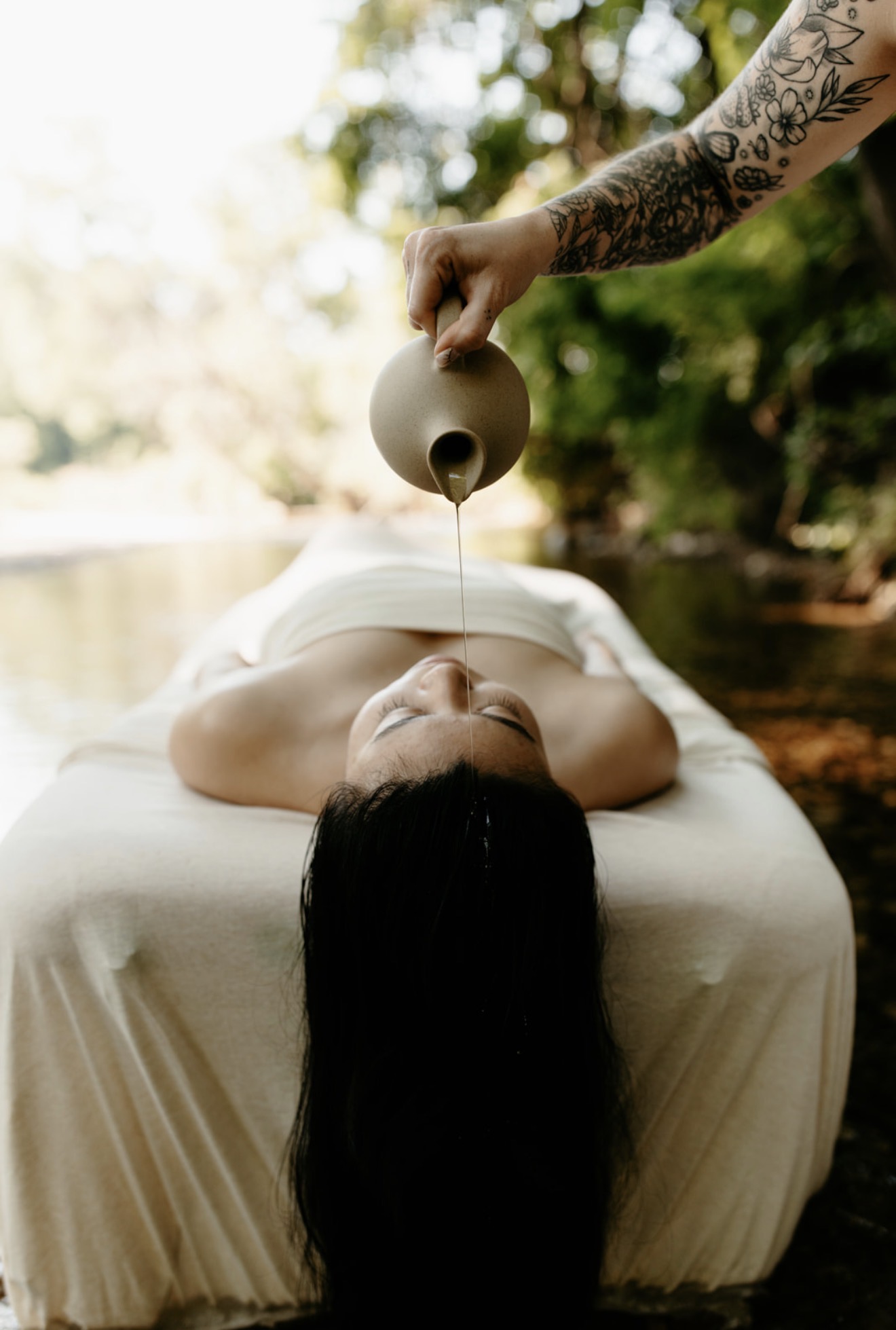 Person lying face up outdoors, receiving a massage with a teapot pouring oil on their forehead, surrounded by trees.