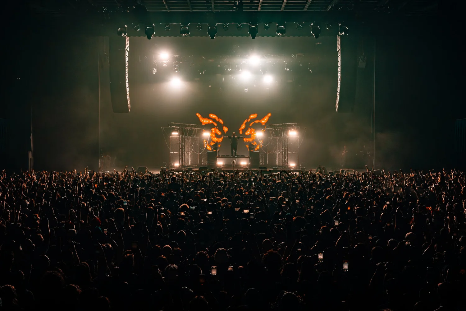 Concert stage with bright lights and flame effects, large audience in front, dark atmosphere.
