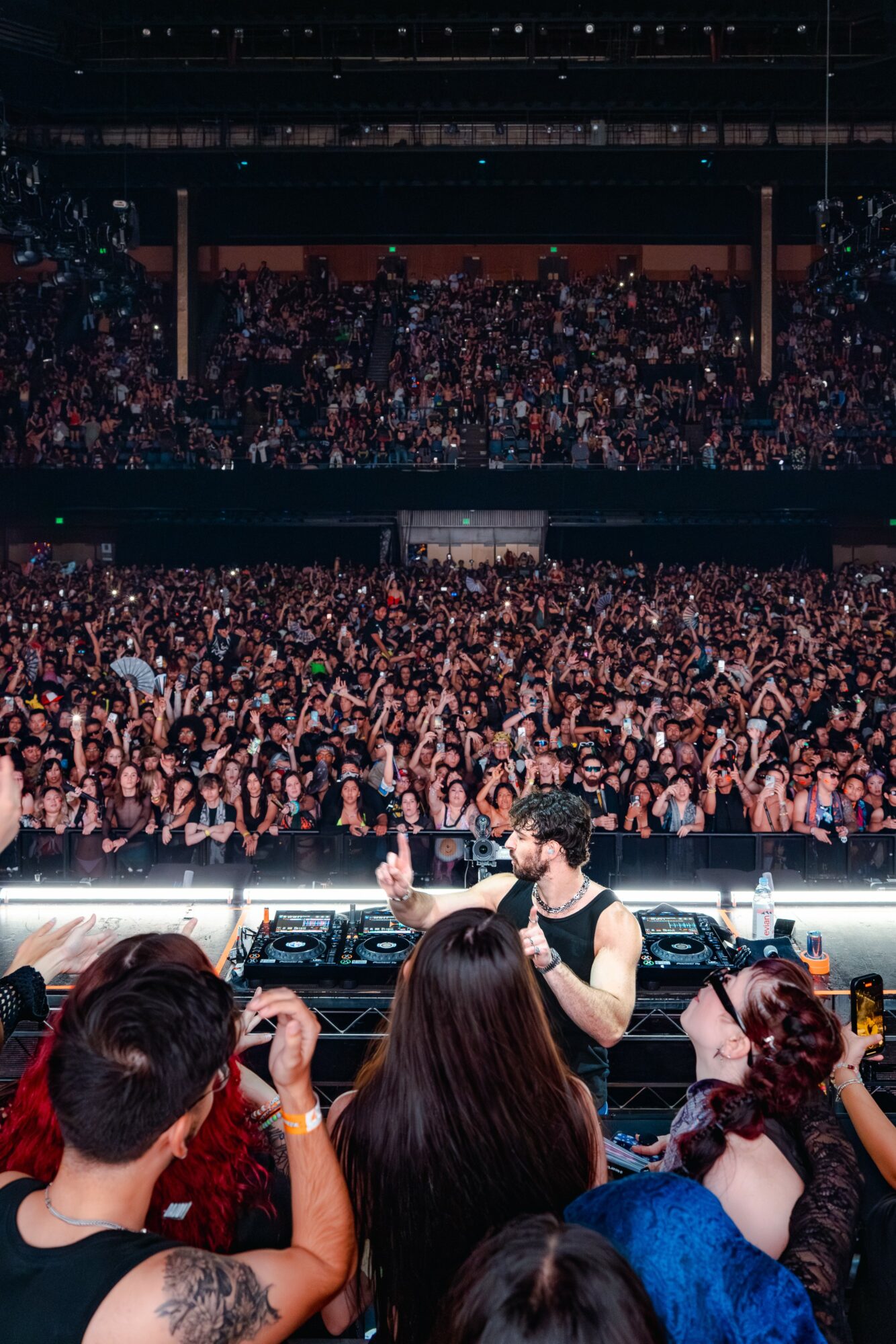 DJ performing on stage with a large crowd in a concert venue, audience raising hands, dark lighting, stage equipment visible.