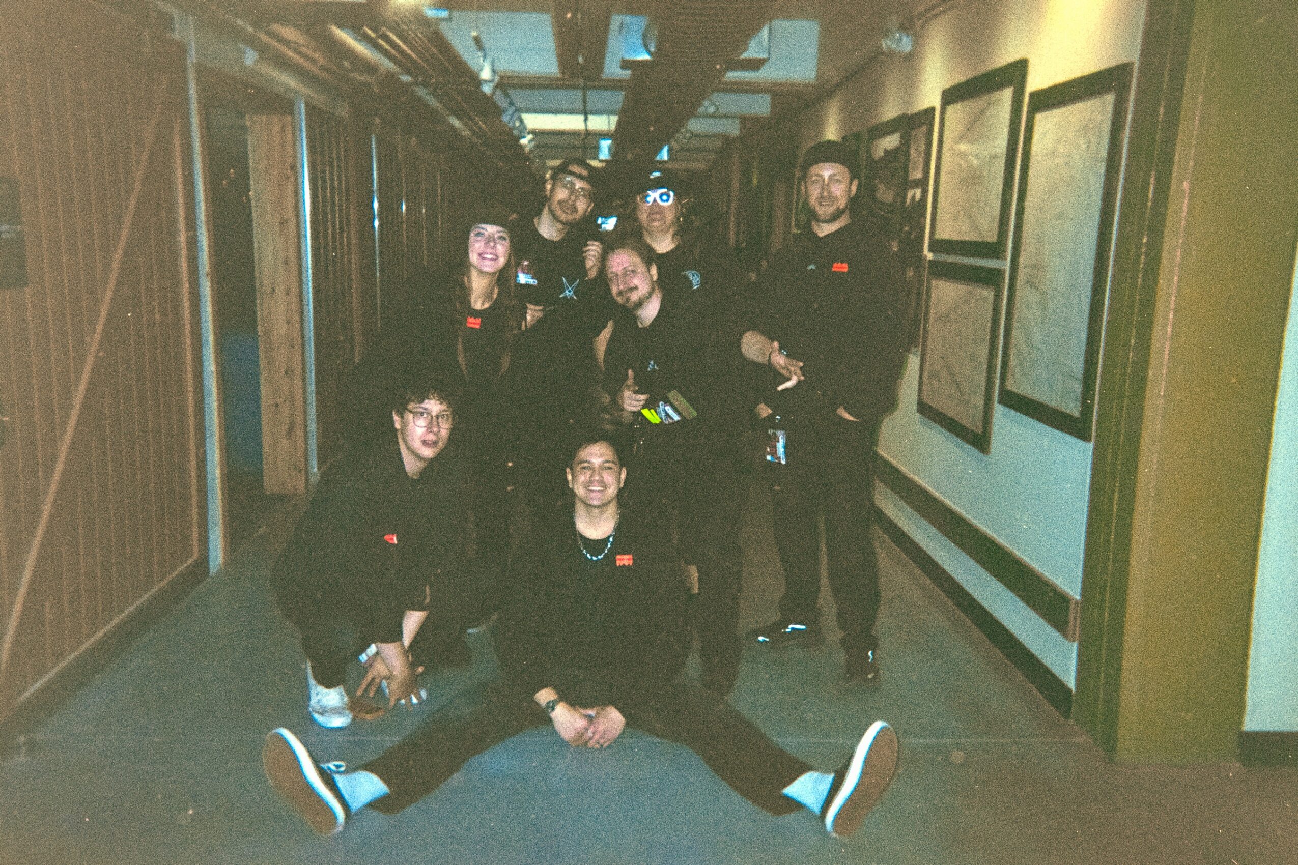 Group of seven people posing in a hallway with framed pictures on the wall, some standing and some sitting on the floor.