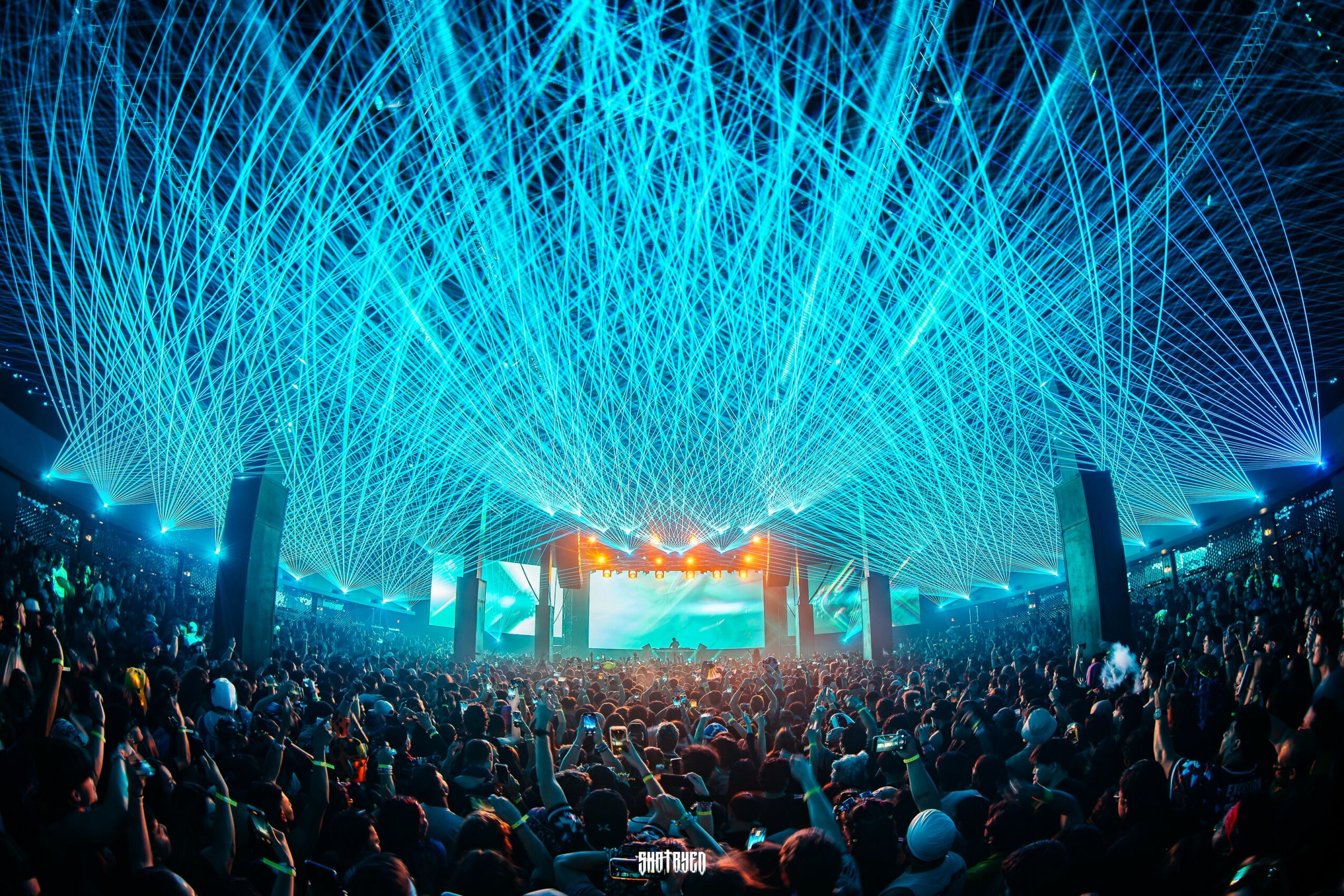 Concert stage with bright blue laser lights and a large crowd, colorful screens, and stage equipment.