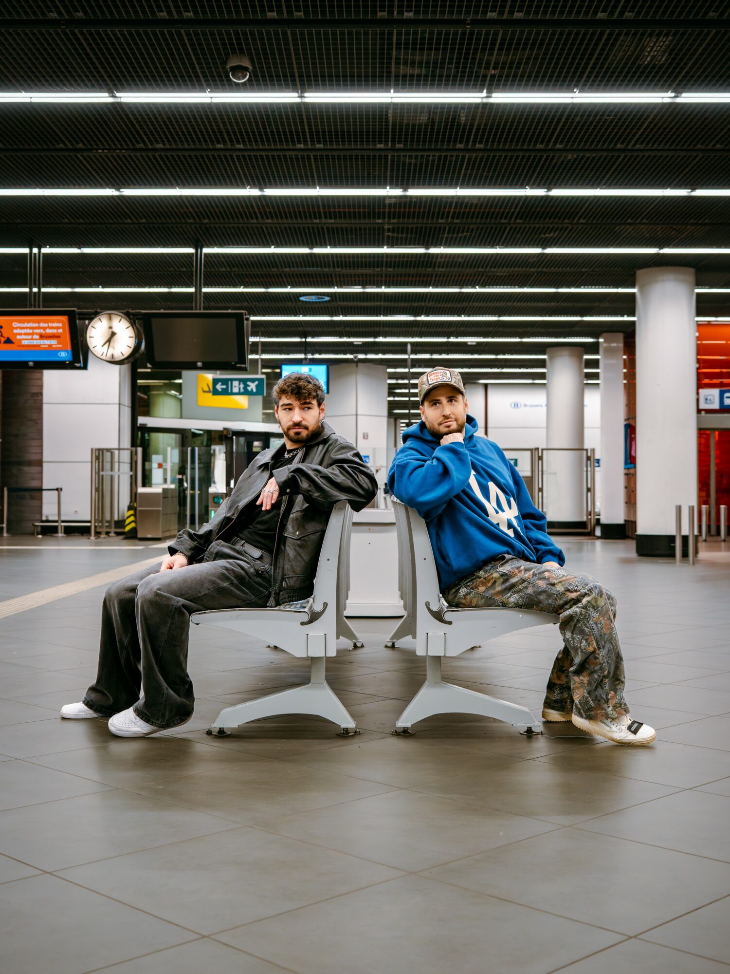 Two men sit on separate chairs facing each other in a modern train station, with a clock and signs overhead.