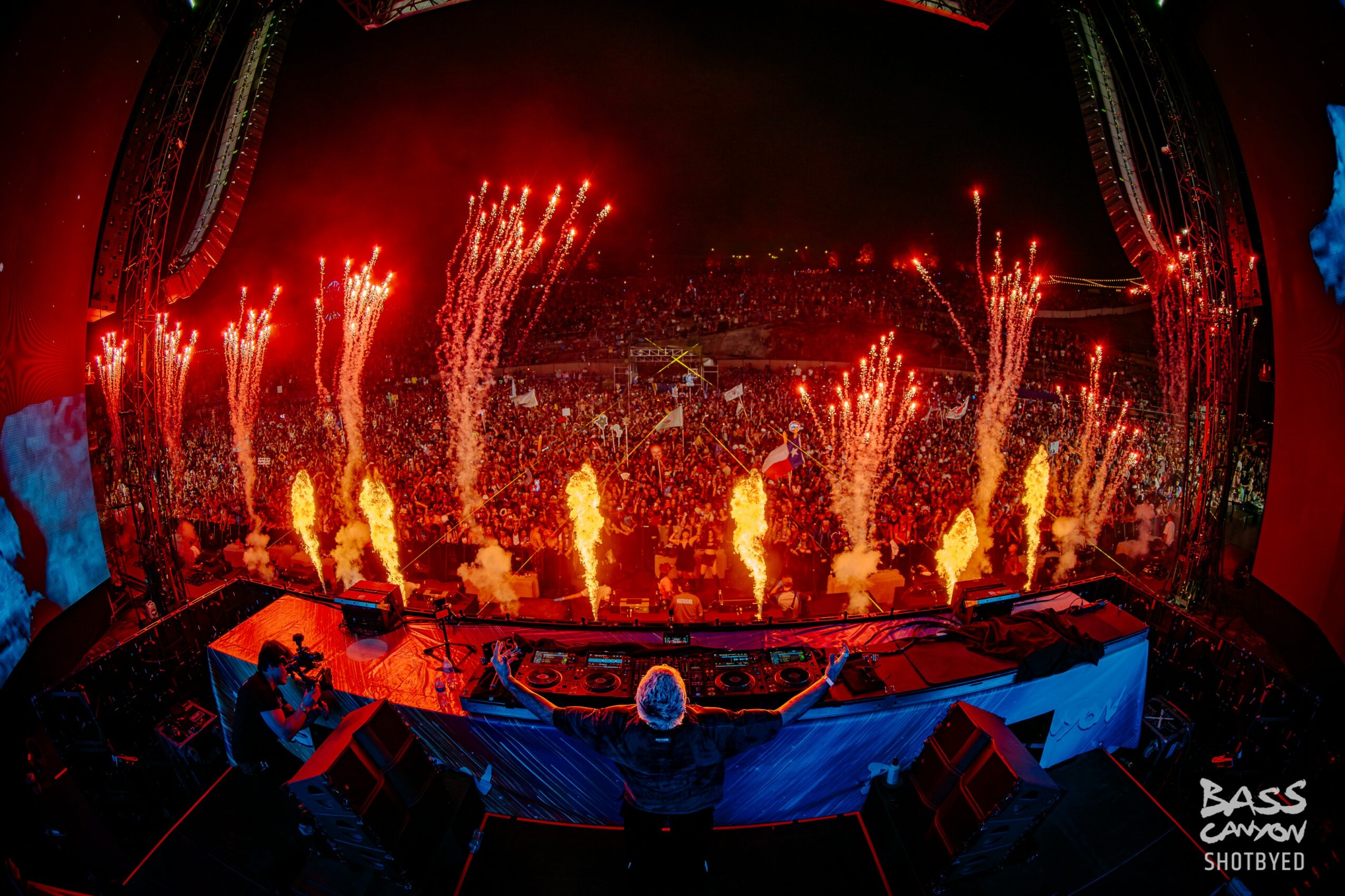 Concert stage with fireworks and pyrotechnics, audience in background, DJ equipment in foreground, vibrant red and orange colors, night scene.