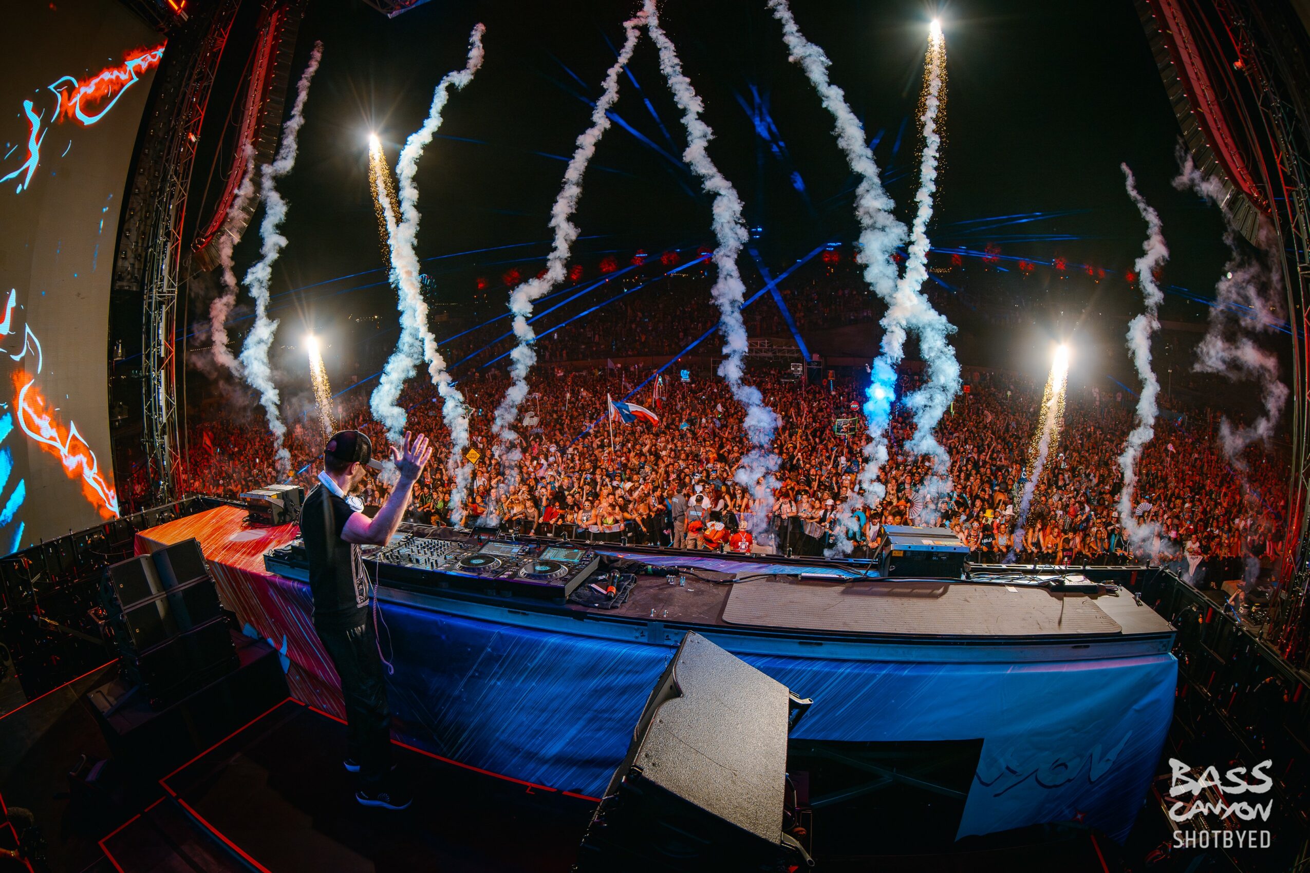 DJ performs on stage with fireworks and a large crowd in the background at a concert.