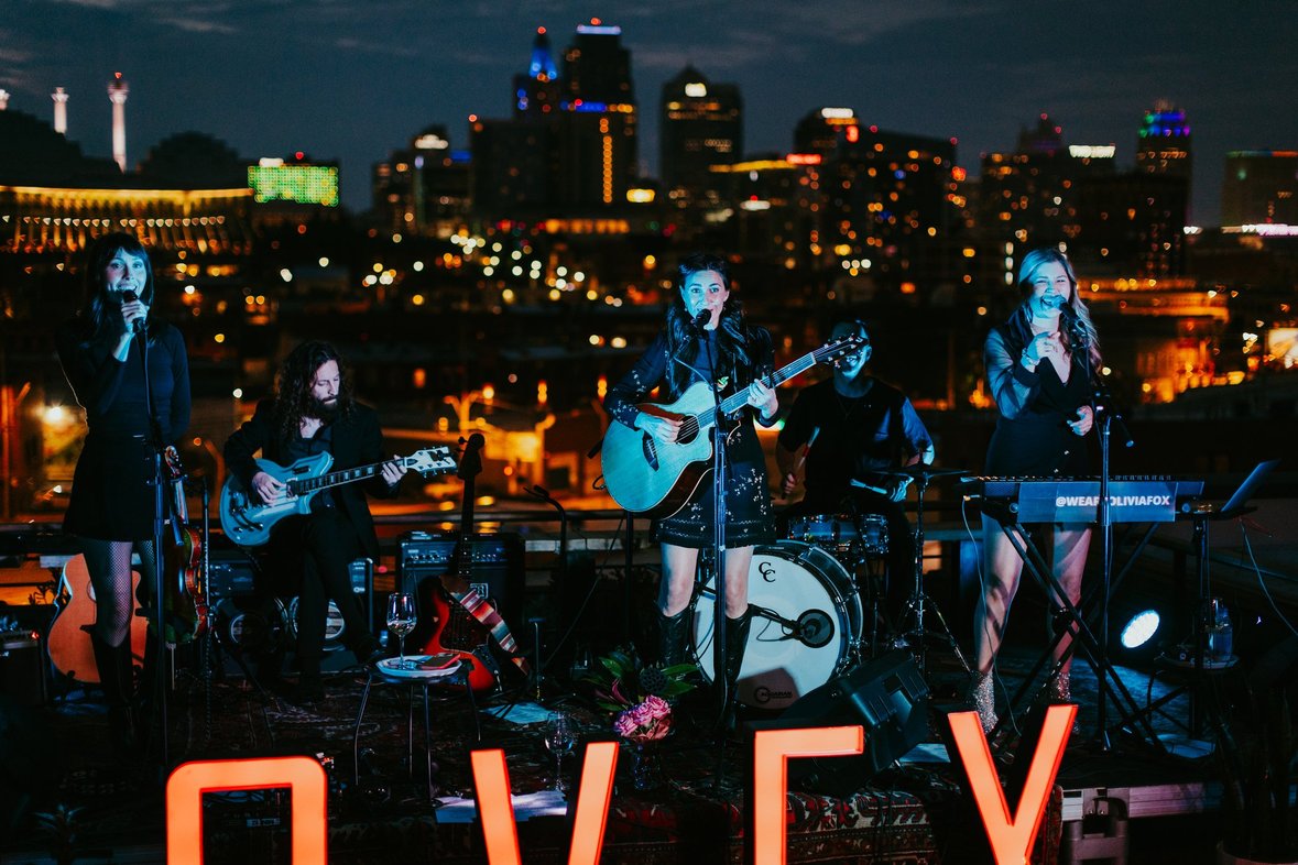 Band performing on rooftop at night with city skyline and lights in background, four women and one man playing instruments.