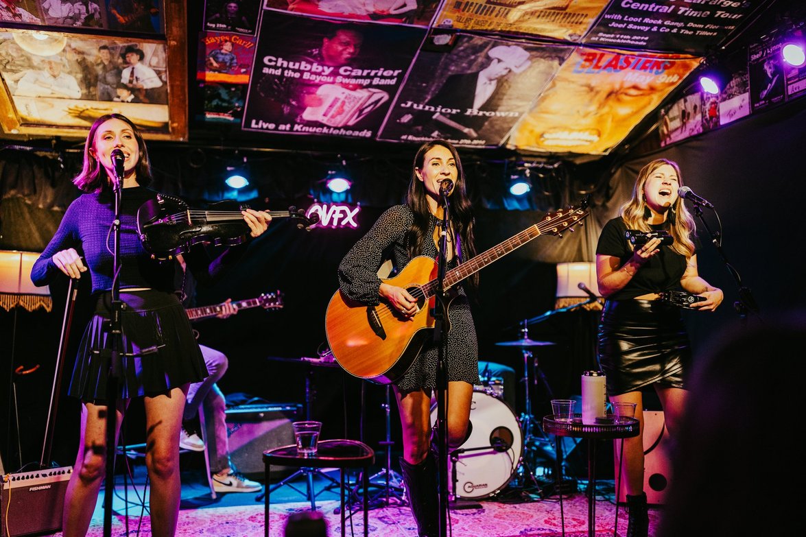 Three women perform on stage with microphones, one playing guitar, in a dimly lit venue with screens above.