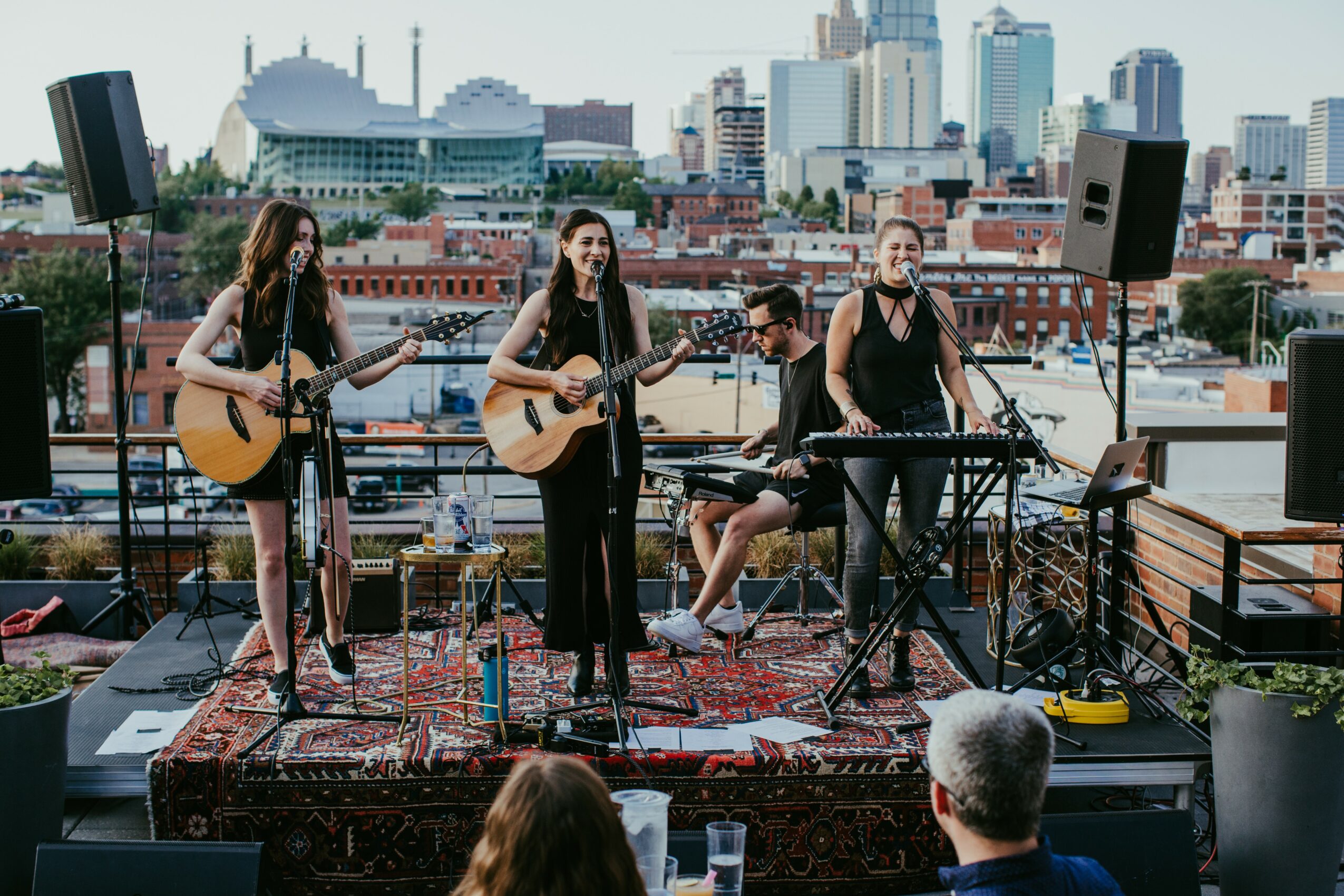 Four musicians perform on a rooftop with city skyline in background, playing guitars, keyboard, and singing, with sound equipment around.