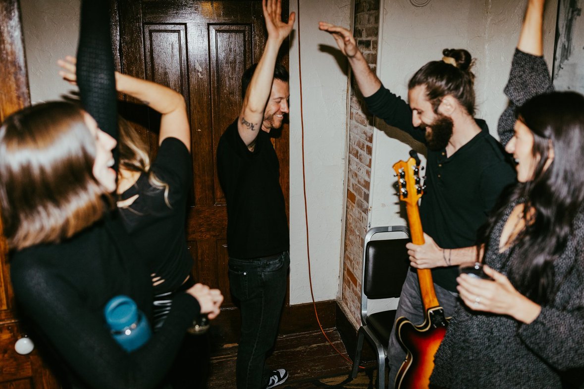 Group of five people dancing and socializing indoors, one holding a guitar, in a cozy room with wooden door and brick wall.