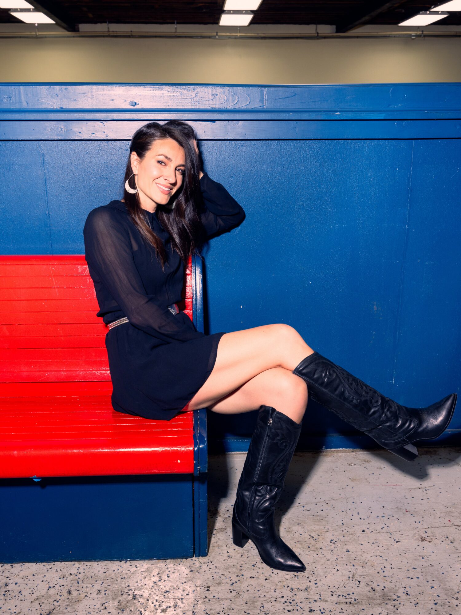 Woman sitting on a red bench, wearing a black dress and knee-high boots, smiling with one hand in her hair.