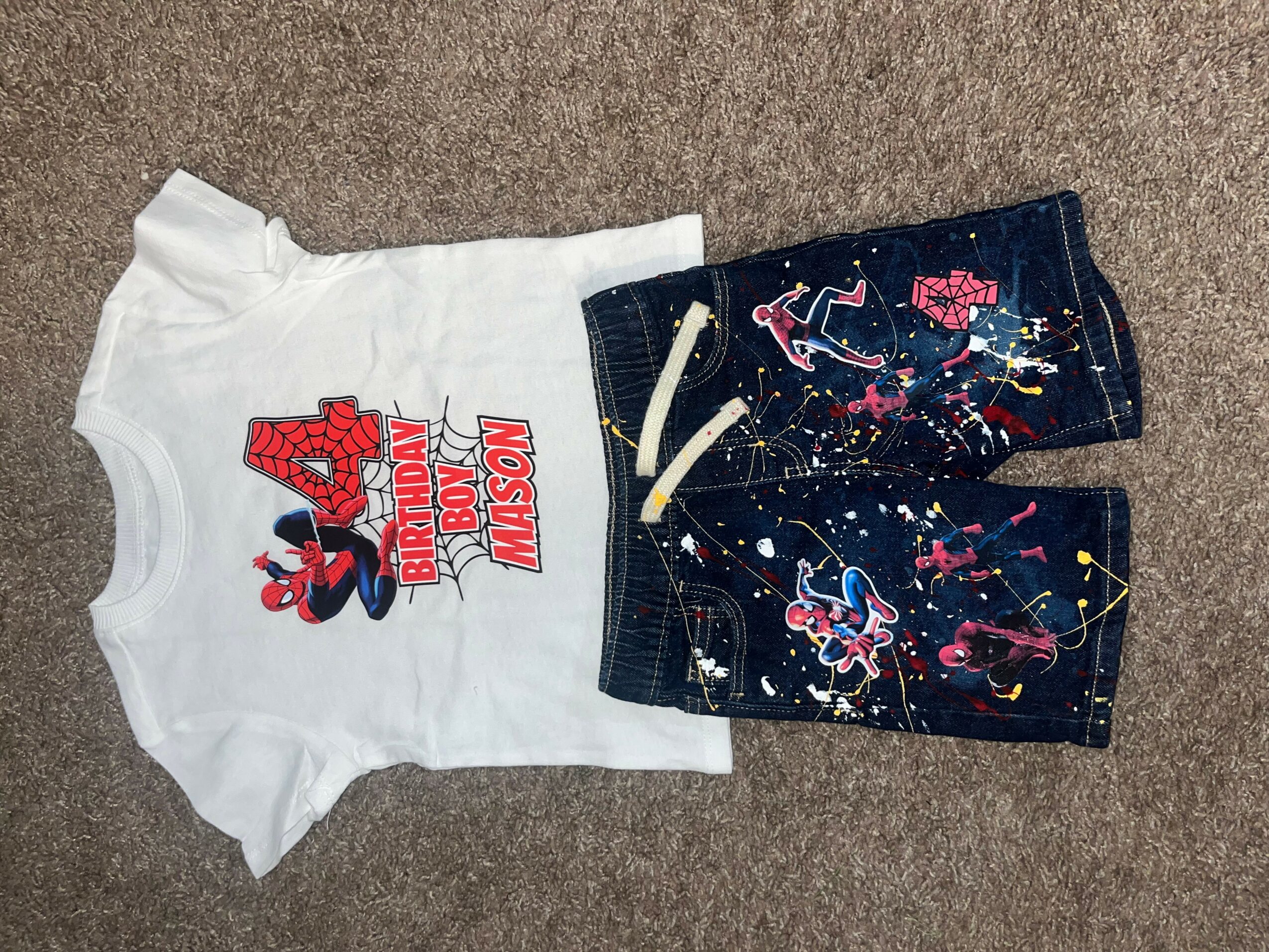 Children's outfit with a white T-shirt featuring Spider-Man and a graphic, paired with dark shorts with colorful patterns, laid on a carpet.