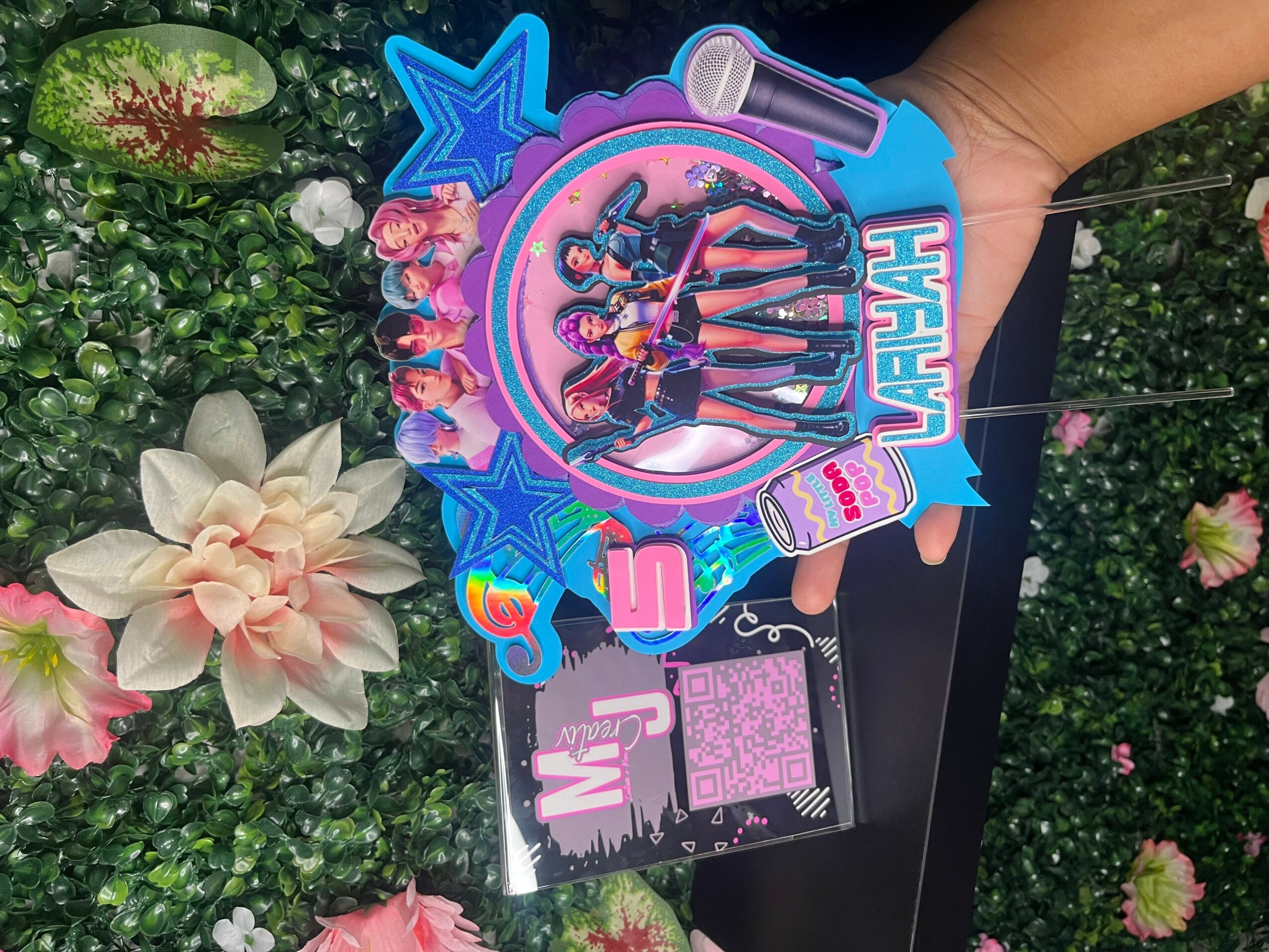 Hand holding a colorful toy with characters, stars, and text, against a background of green plants and flowers.
