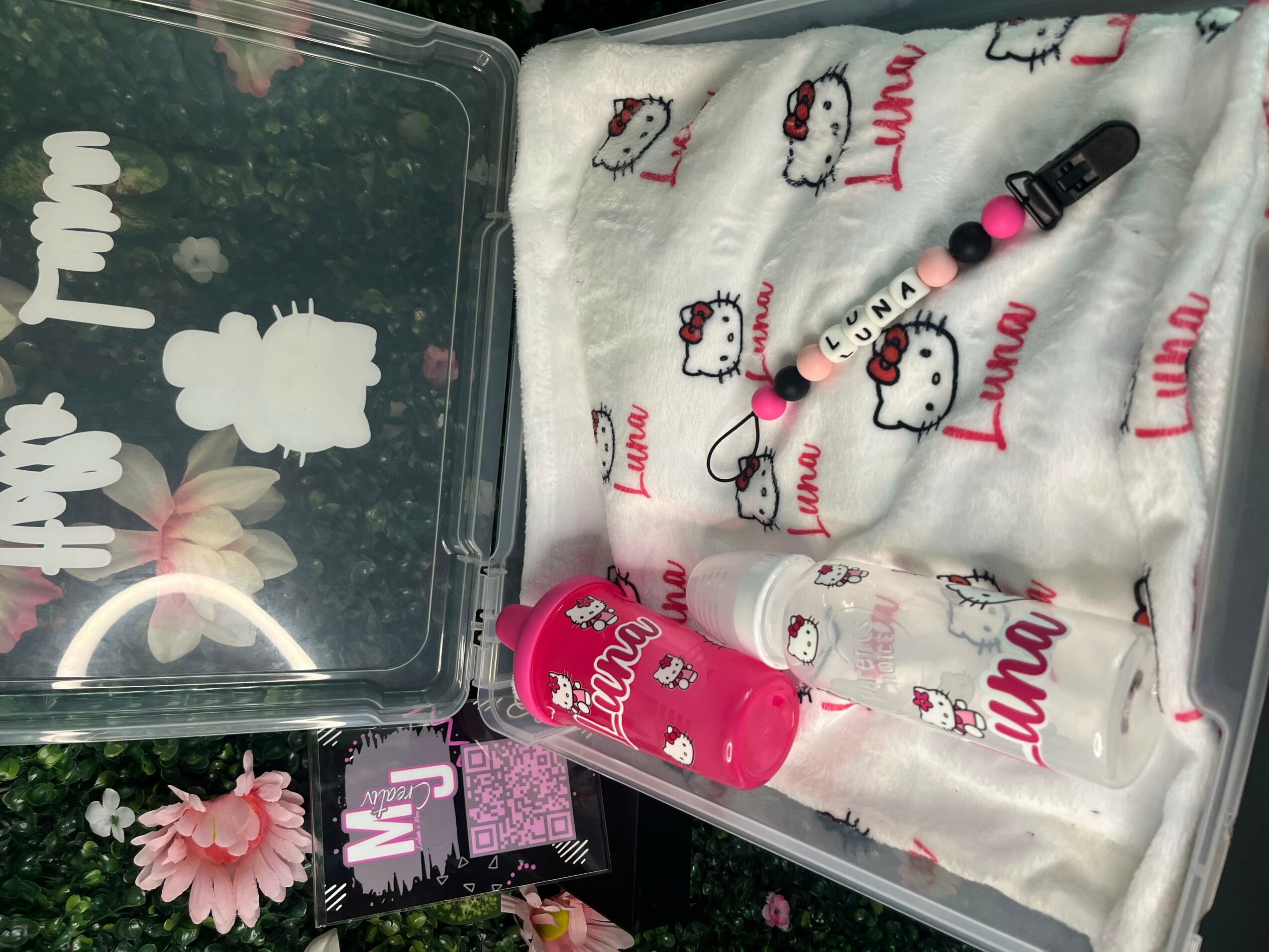 A Hello Kitty-themed cloth, a pink lipstick, and a black and pink beaded bracelet on a white fabric with Hello Kitty and 'Luna' text, with a plastic container nearby.