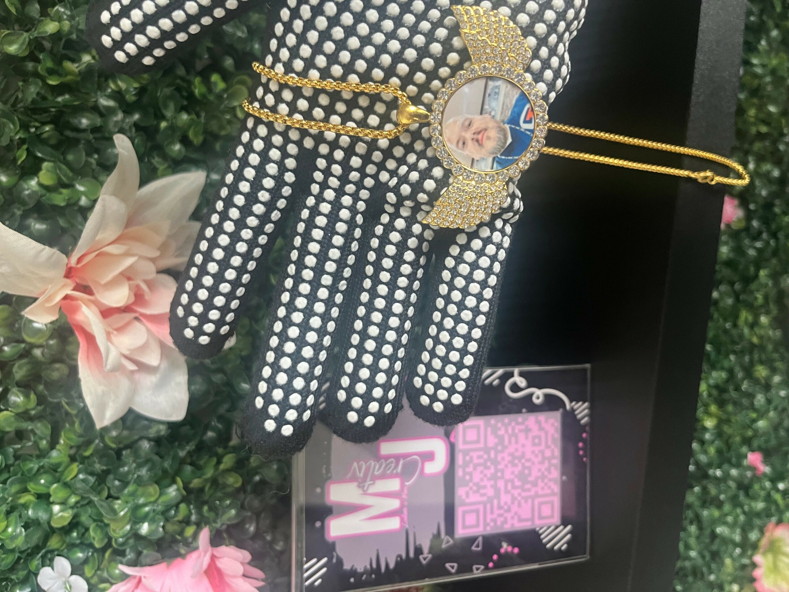 Glove-shaped jewelry with a portrait, surrounded by rhinestones, displayed outdoors with flowers and greenery.