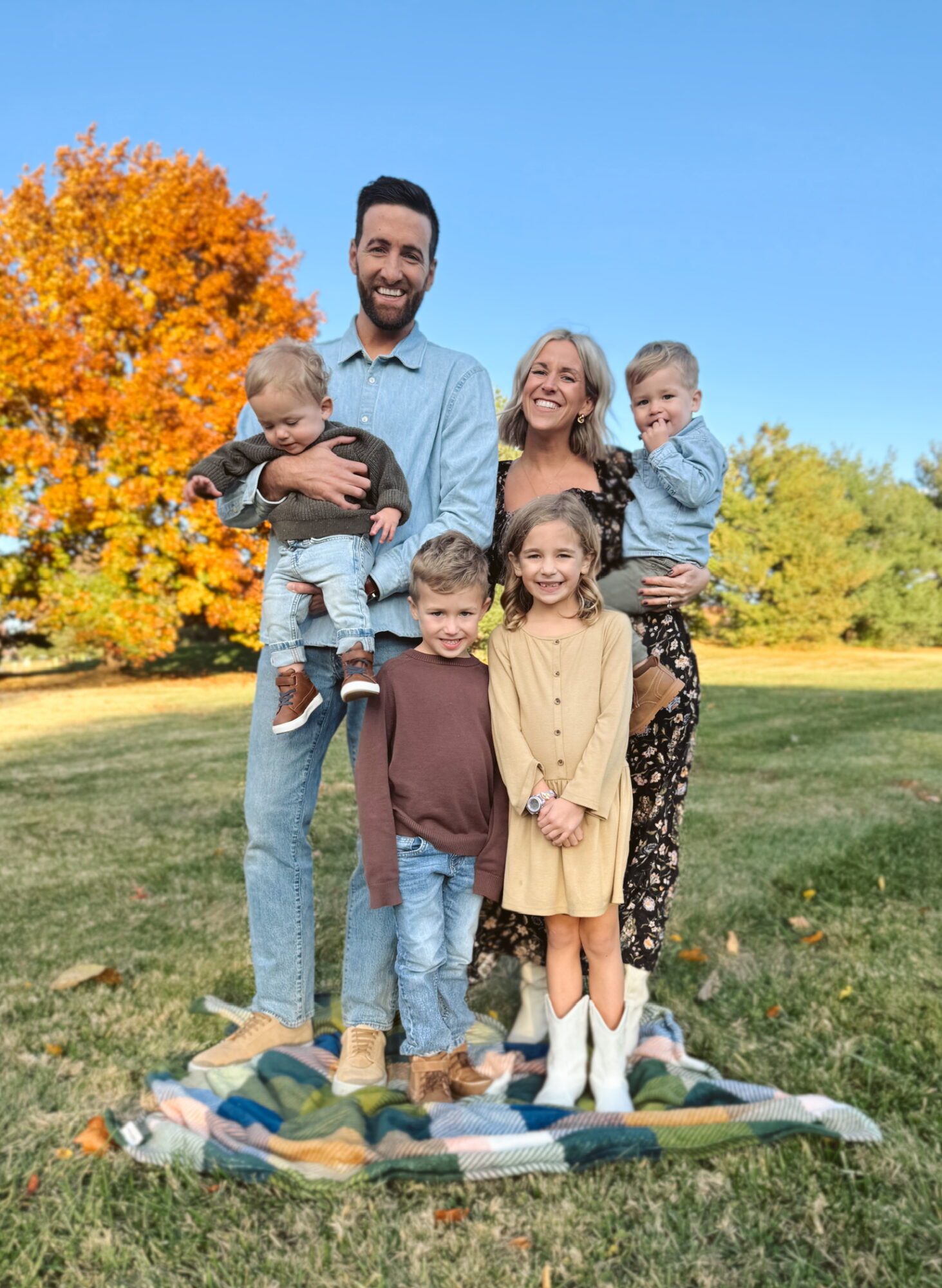 Family of six standing outdoors on a blanket, smiling, with trees and blue sky in background.