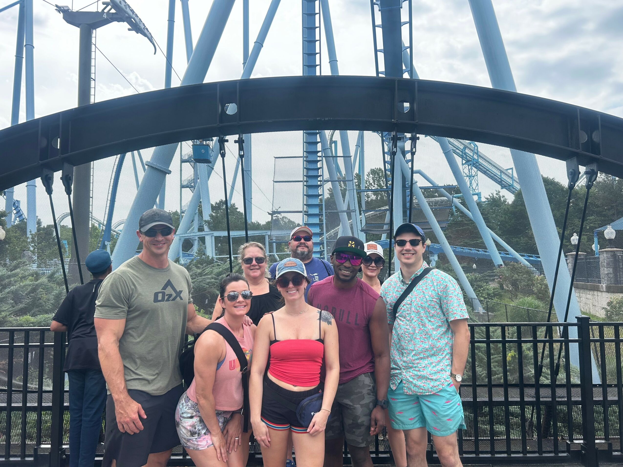 Group of seven people standing in front of a roller coaster at an amusement park.