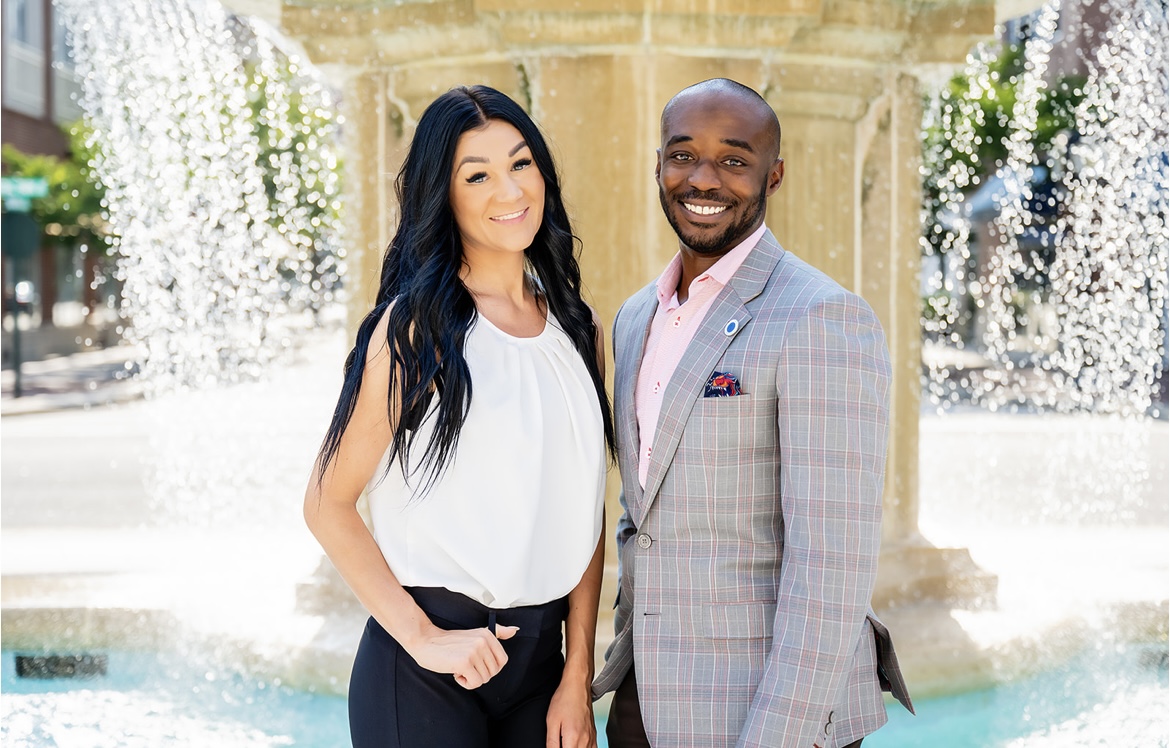 A smiling woman and man stand in front of a fountain with water streams, dressed in formal attire, outdoors.