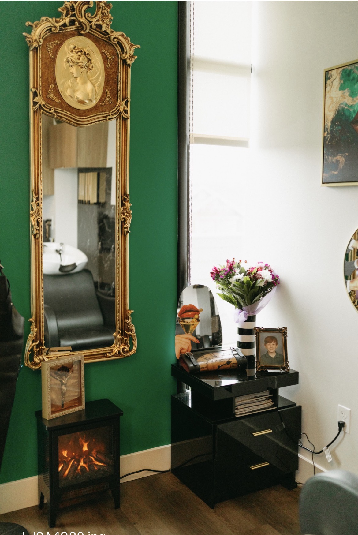 Decorative mirror with gold frame on green wall, small black table with flowers and framed photo, white wall with window, wooden floor.
