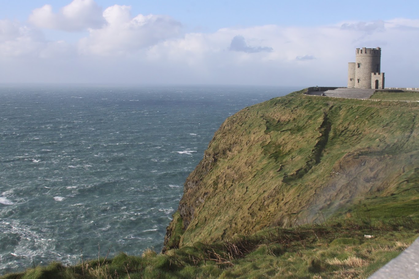 Cliffside with a tower overlooking the ocean, cloudy sky, grassy terrain, and waves crashing below.