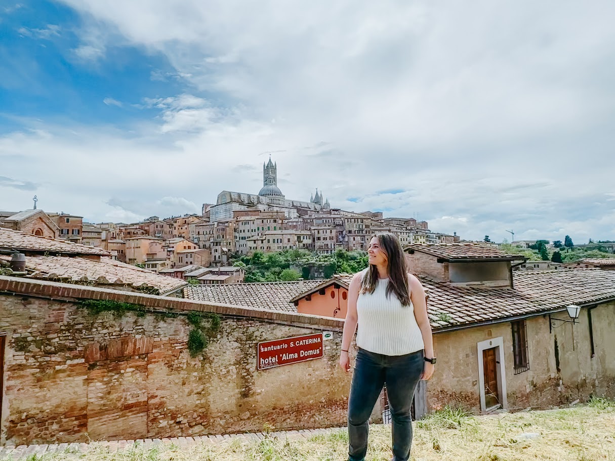 Woman standing outdoors with a cityscape and a large church or cathedral in the background, cloudy sky, stone wall in foreground.