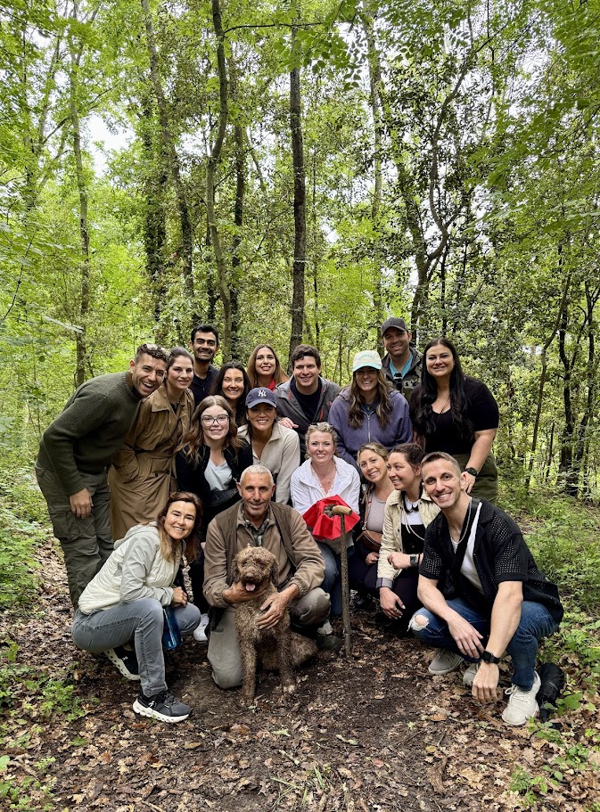 Group of people outdoors in a forest, smiling, with a dog, some wearing hats, surrounded by trees and greenery.
