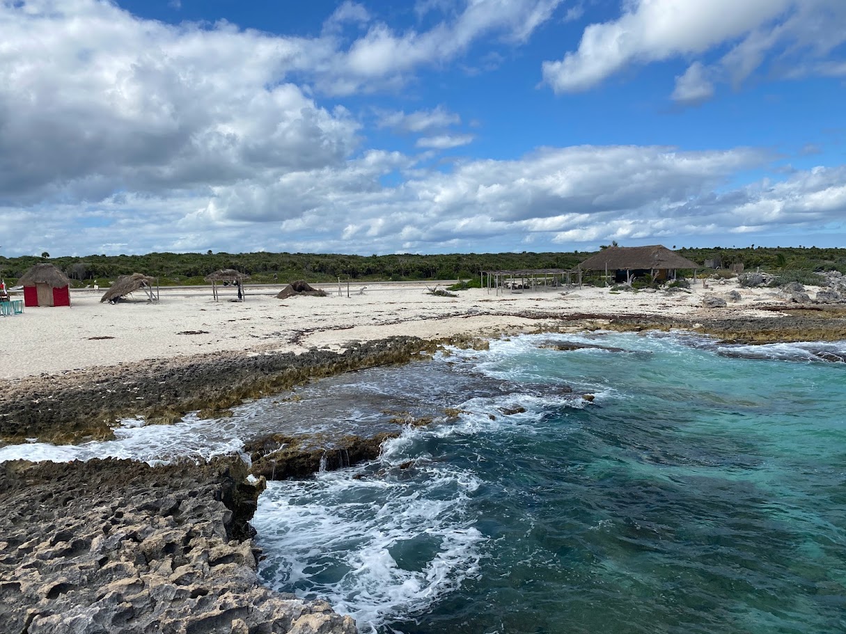 Beach with sandy shore, rocks, and turquoise water under a partly cloudy sky, with a small building in the background.