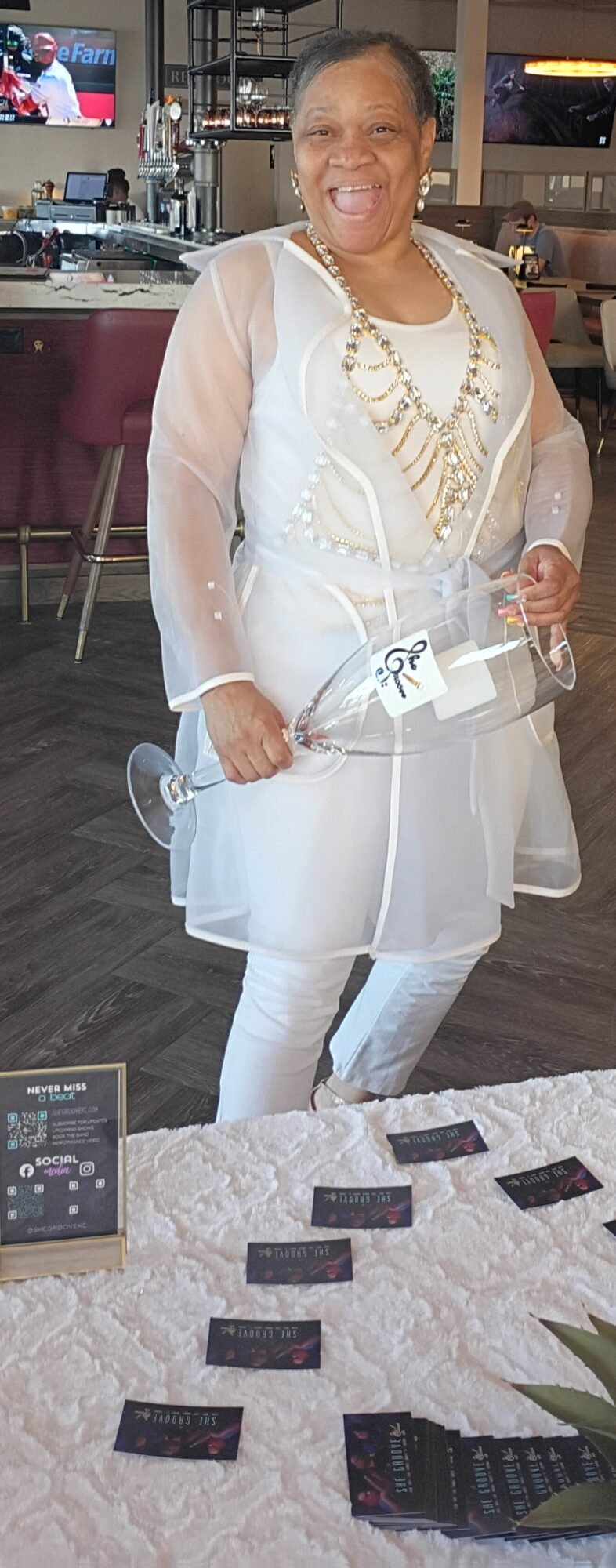Woman smiling, dressed in white, standing indoors with a table of cards in front, holding a clear clutch, background of a restaurant.