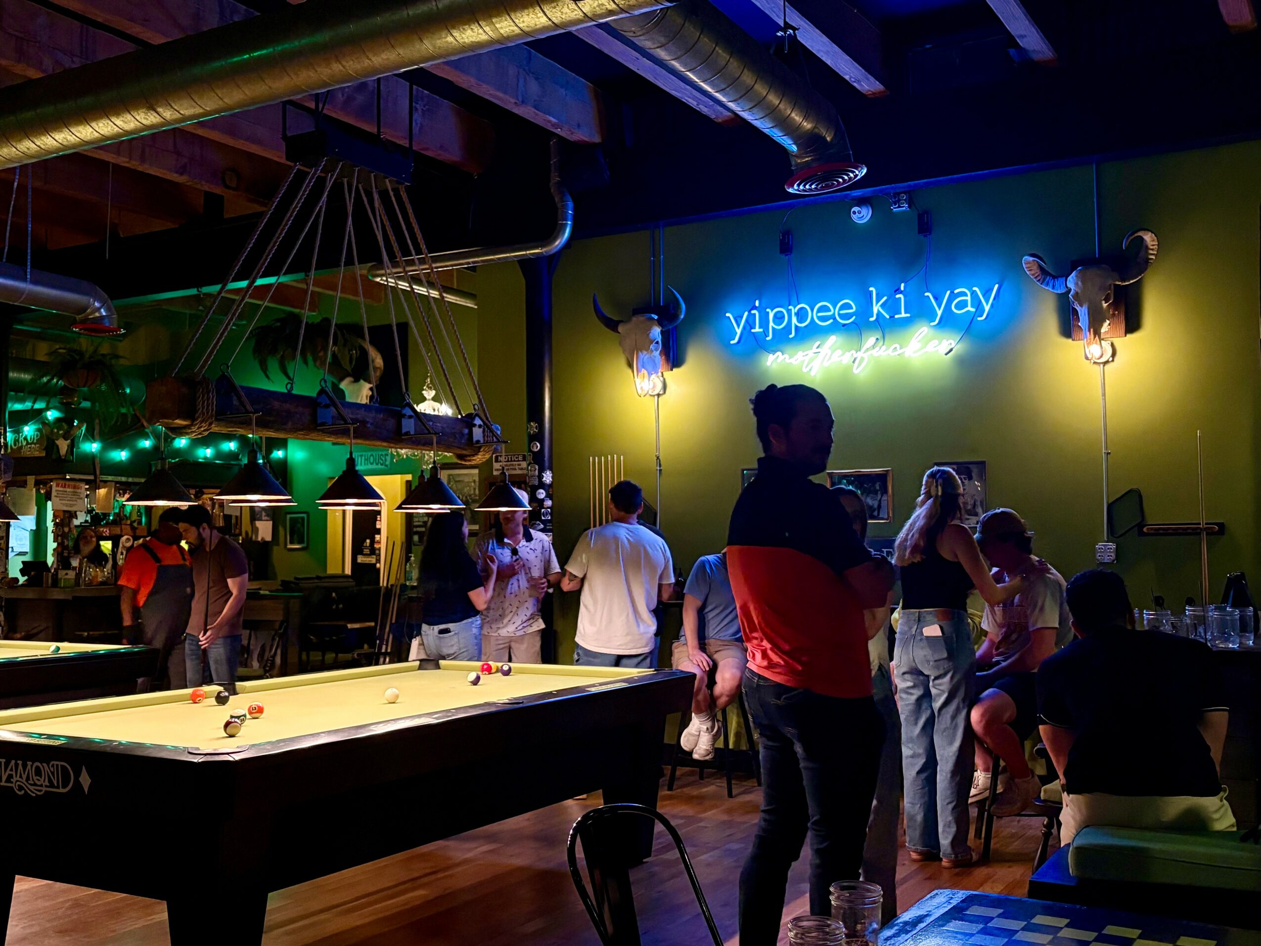 People gather in a bar with a neon sign, pool table, and decorative lighting, enjoying social activities.