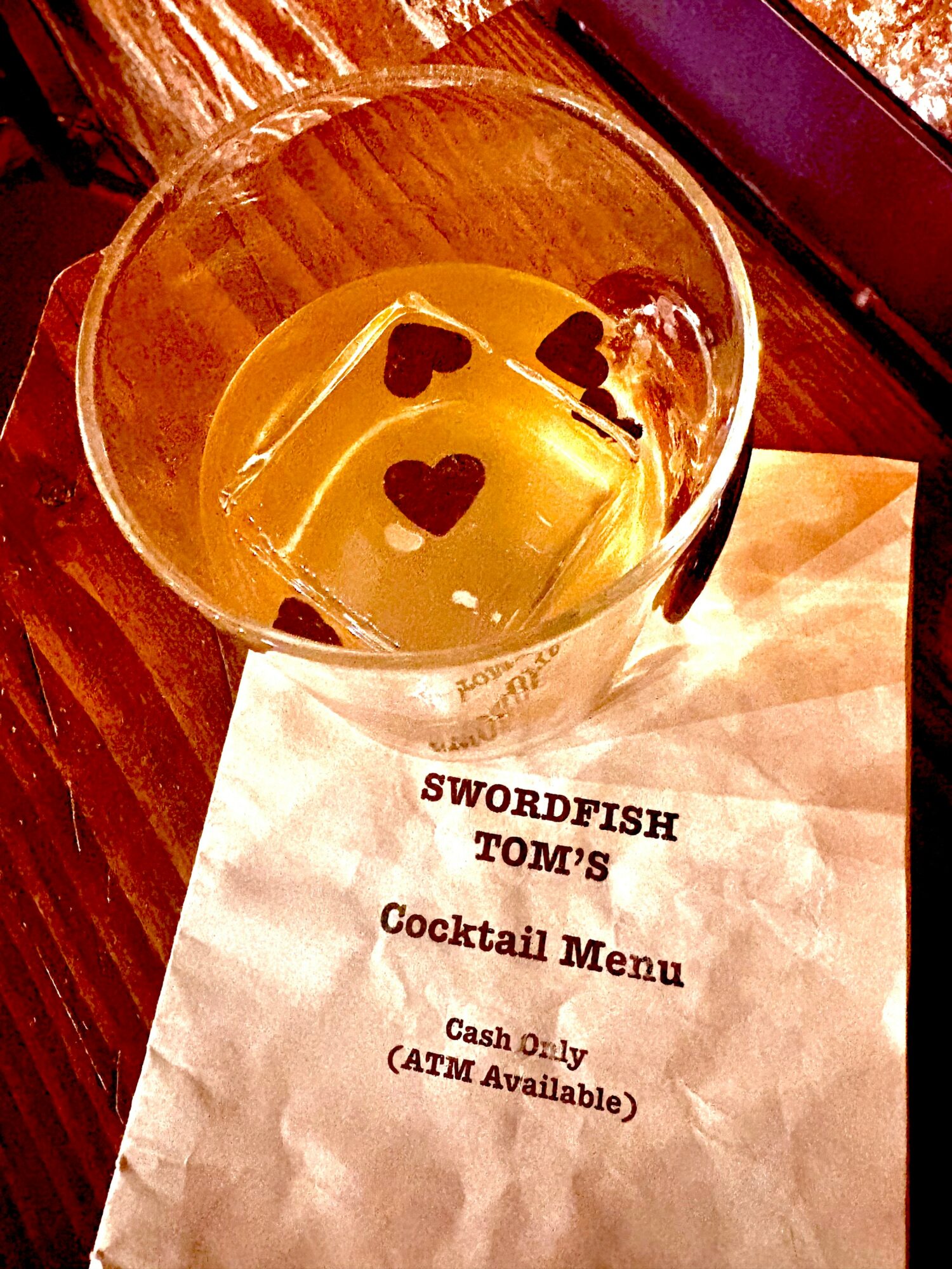 A glass with a drink and heart-shaped ice cubes on a table, with a paper menu underneath.