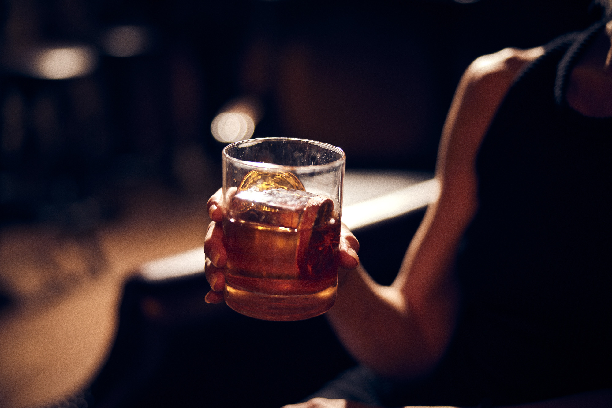 Person holding a glass of dark-colored beverage with ice, in a dimly lit setting.