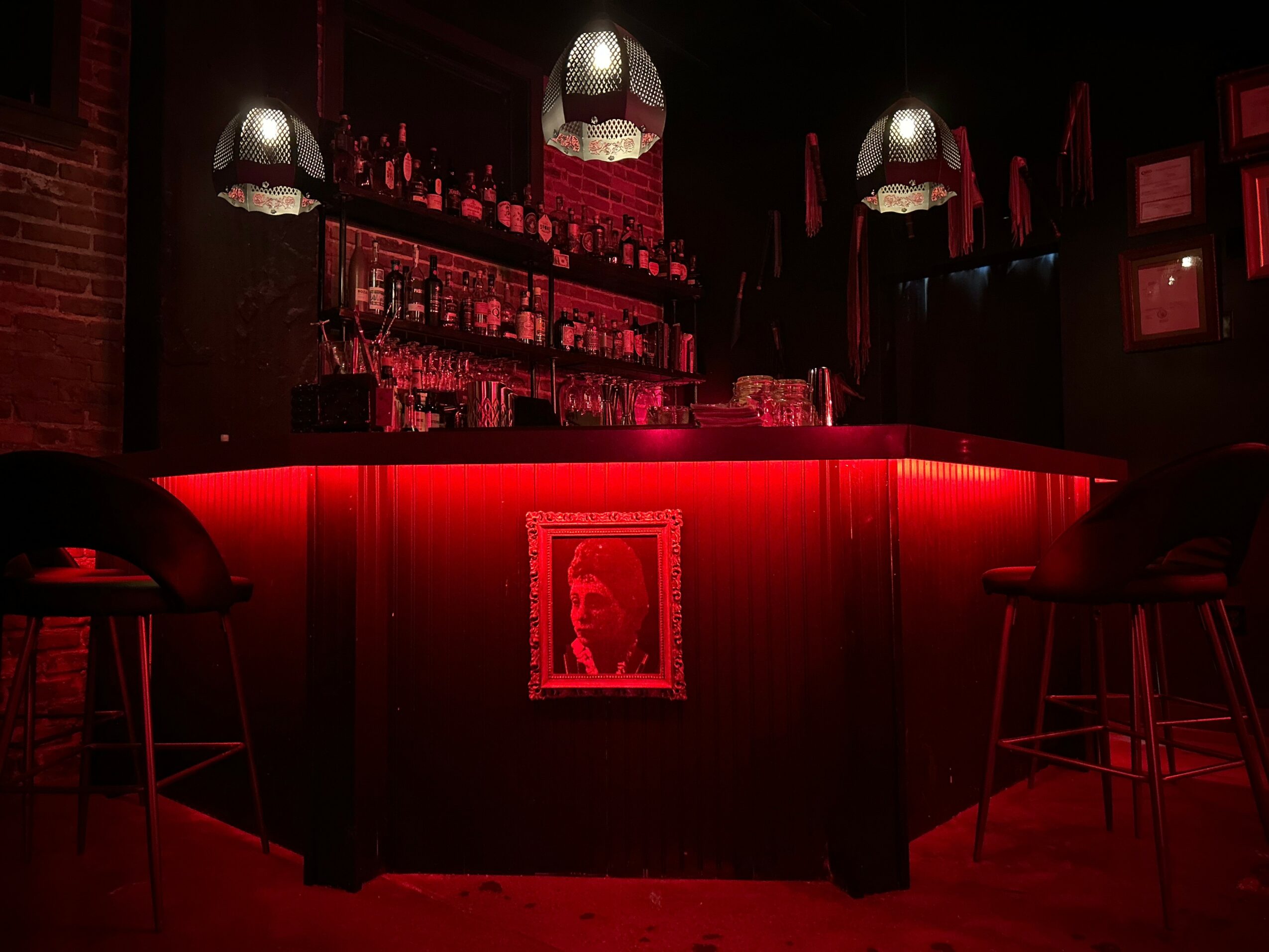Bar with red lighting, shelves with bottles, framed picture on front, three hanging lamps, two stools on each side.