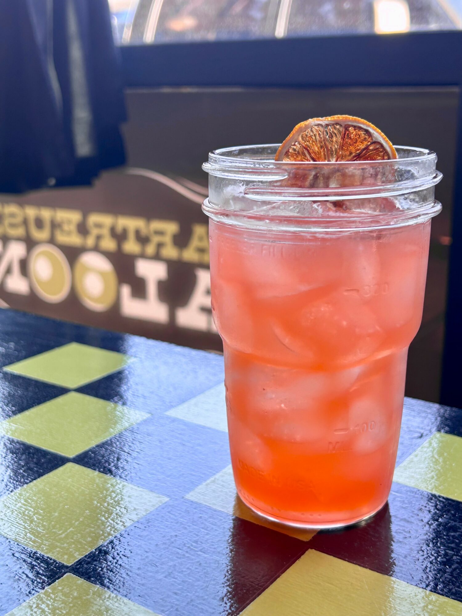 A pink-colored drink with a dried citrus slice on top, in a clear plastic cup on a checkered table.
