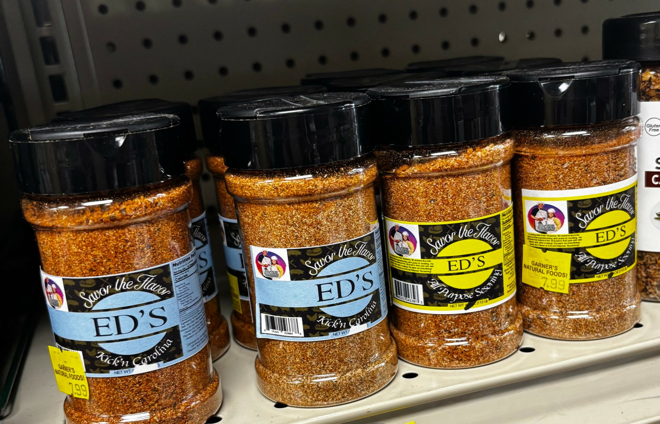Multiple jars of seasoning on store shelf, black lids, labels with blue and yellow accents, brand name ED'S, in a retail setting.