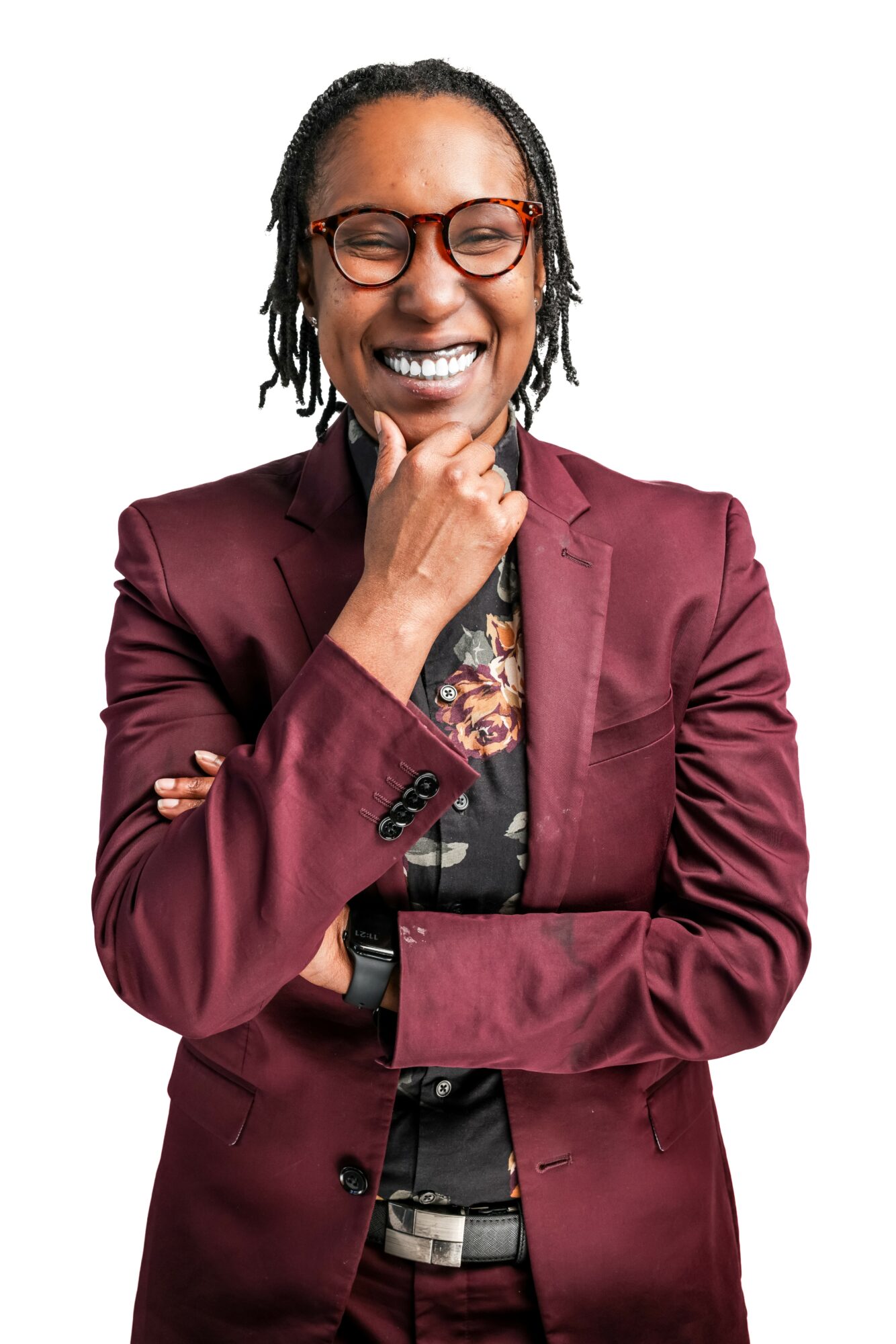 Smiling woman with glasses, wearing a maroon blazer and floral shirt, with hand on chin, against white background.