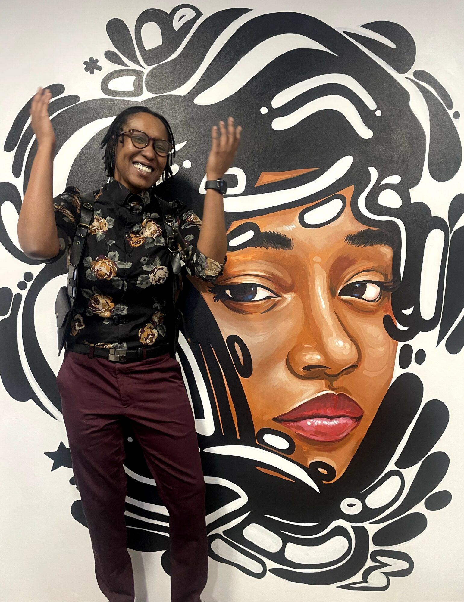Person smiling and raising hands in front of a large black and white mural of a woman's face with detailed hair and facial features.