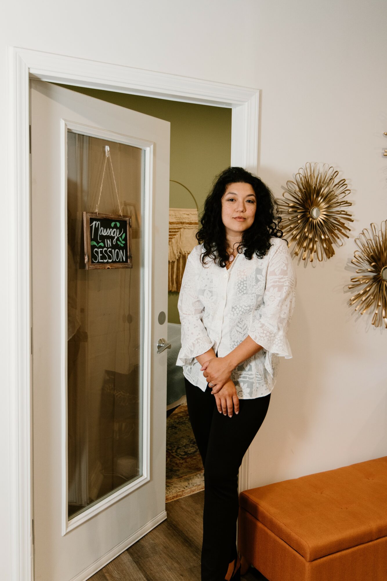 Woman with dark curly hair in white blouse and black pants standing indoors near a door with a sign, decorated wall, and orange bench.