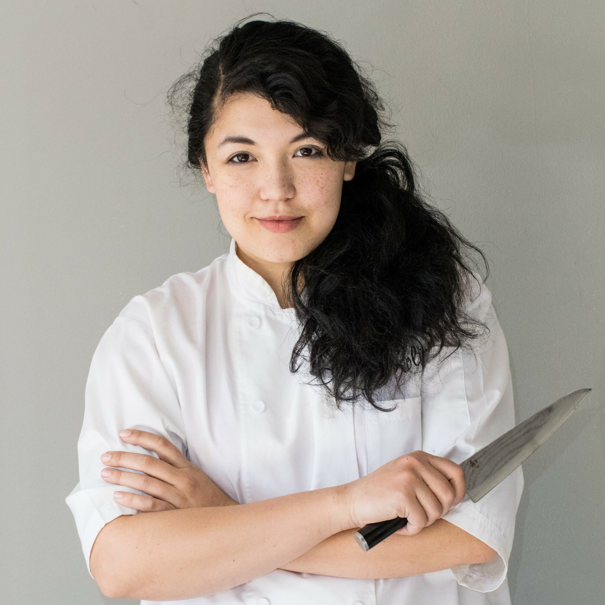 Young woman with dark, curly hair wearing a white chef's coat, holding a clipboard, smiling softly, against a plain background.