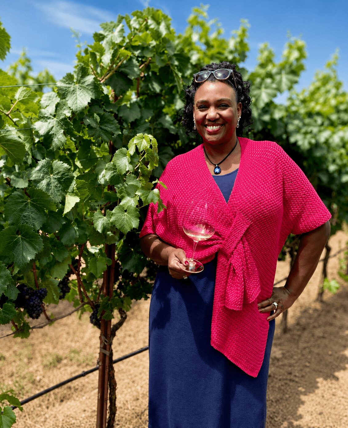 Woman in pink sweater holding a glass of white wine outdoors with green grapevines and blue sky behind her.