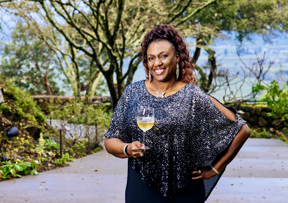 Woman smiling outdoors holding a glass of white wine, trees and pathway in background.