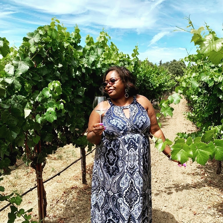 Woman in sunglasses holding a glass of wine in a vineyard with green vines and blue sky