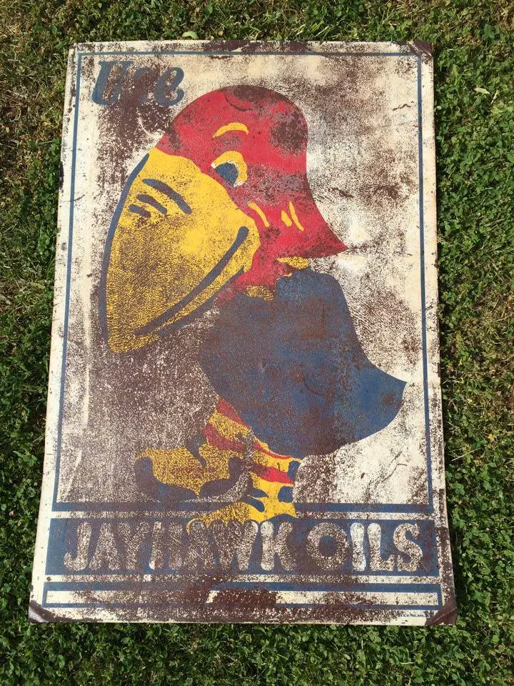 Weathered sign with a cartoon duck wearing a red cap and blue outfit, advertising Jayhawk Oils, on grass.