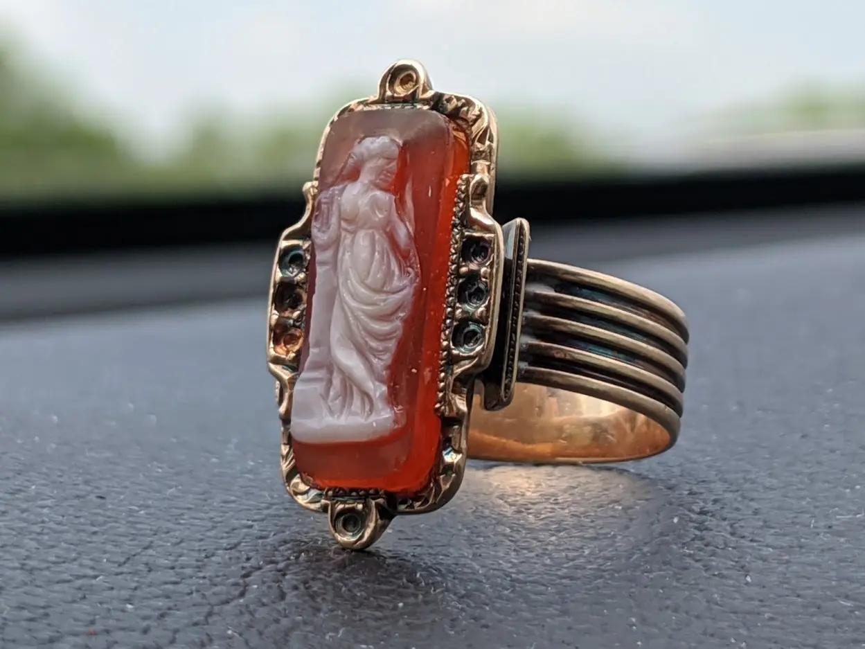 Ring with a rectangular carved stone set in a decorative metal band, placed on a dark surface.