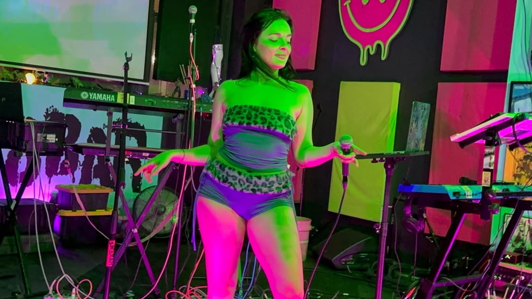 Woman on stage with musical equipment, wearing glasses and colorful clothing, holding a microphone in each hand.