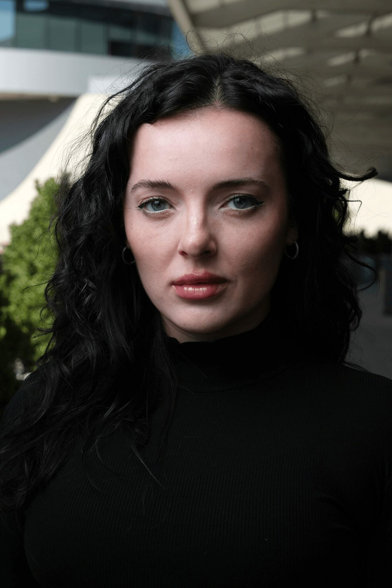 Young woman with dark wavy hair and light makeup, wearing a black top, outdoors with modern building background.
